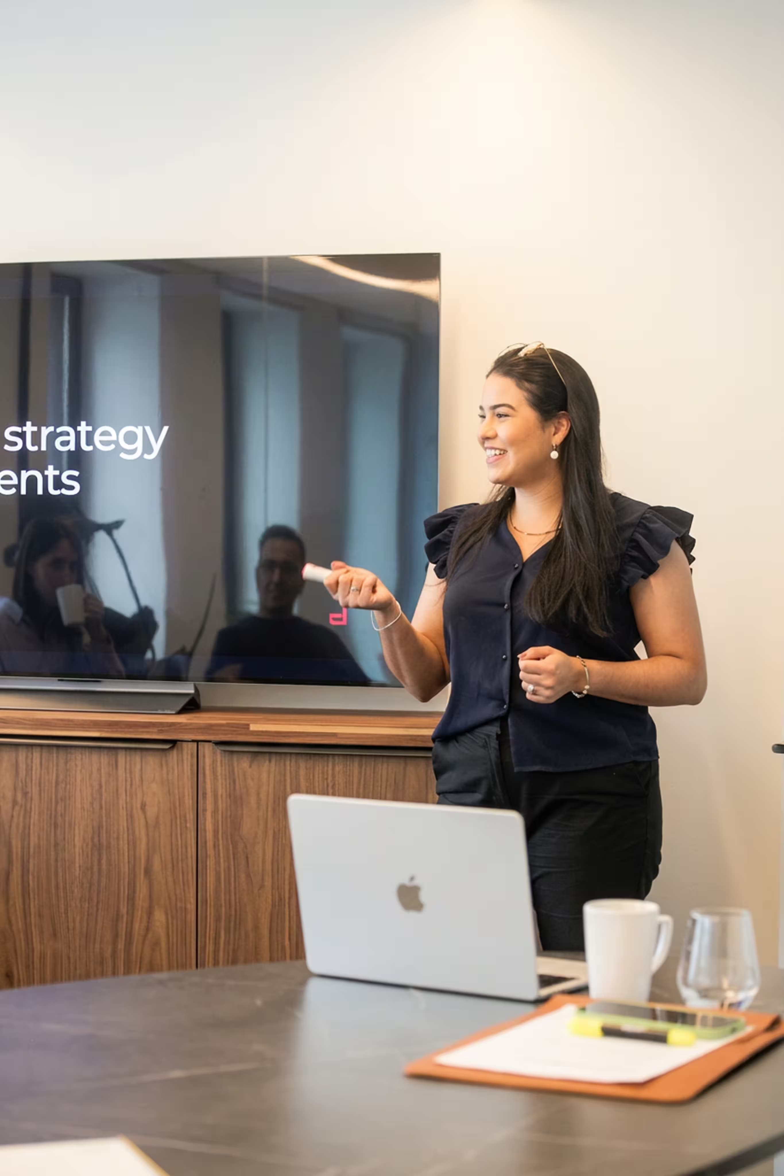 Manoela Bezerra presents in a conference room beside a laptop and large display.